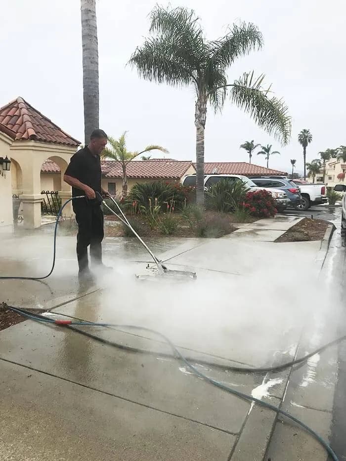 Pressure washing a walkway with heavy spray, palm trees and a tile roof behind